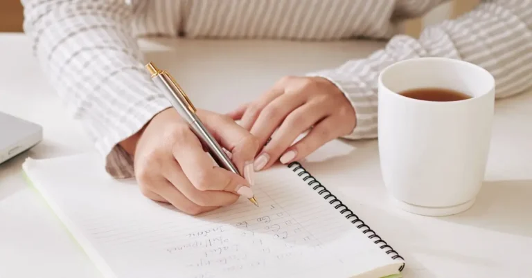 how to write affirmations + A person sits at the desk, writing affirmations in their journal and drinking coffee